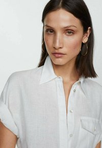 Camicia bianca in lino con colletto a pulsante e maniche corte arrotolate; presenta una tasca frontale e bottoni perlati.