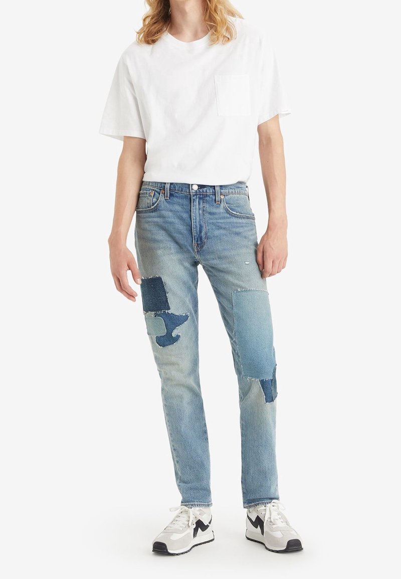 Jeans en denim bleu clair avec des détails en patchwork dans des teintes plus foncées, portés avec un T-shirt blanc uni et des baskets noir et blanc.