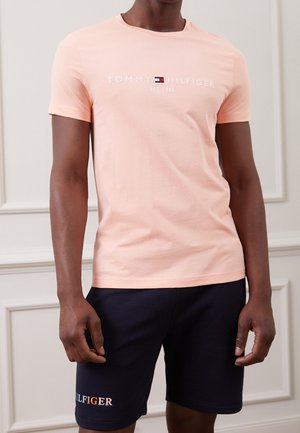 Różowa bawełniana koszulka z okrągłym dekoltem i krótkimi rękawami, z logo "TOMMY HILFIGER" w kolorze białym. Noszona z granatowymi szortami, które mają logo w kolorze pomarańczowym.