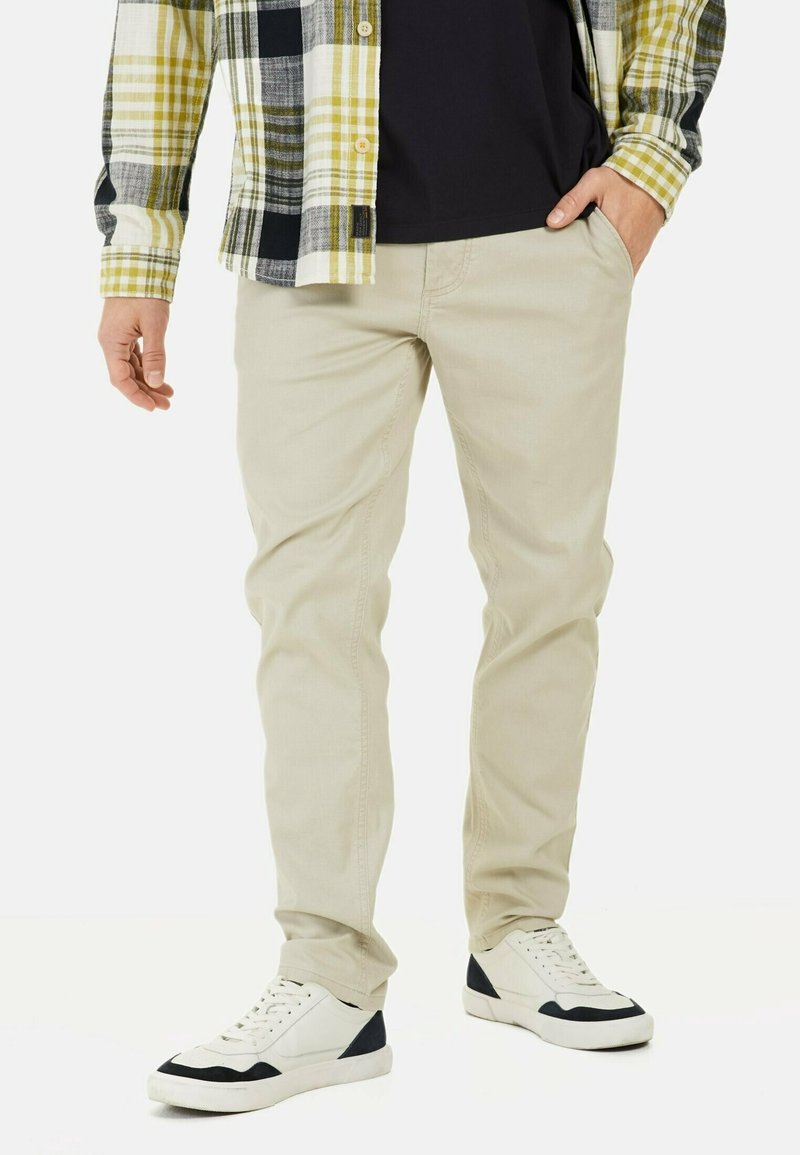 Pantaloni in cotone beige con una vestibilità slim, indossati con una maglietta nera e una camicia a scacchi con dettagli verdi e gialli. Sneakers bianche.