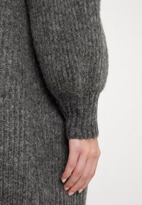 Detail eines grauen Strickpullovers mit geripptem Muster und auffälligem Puffärmel. Eine sichtbare Hand liegt auf dem Stoff und hebt dessen Material hervor.