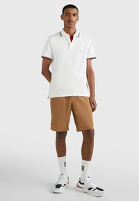 Polo shirt bianca con rifiniture rosse e blu navy, abbinato a pantaloni corti beige. Il modello indossa sneakers bianche e calze fino al ginocchio con un logo.