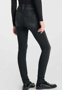 Jean skinny en denim noir avec une taille haute, comportant deux poches arrière et des détails de couture subtils. Portés avec des chaussures noires.