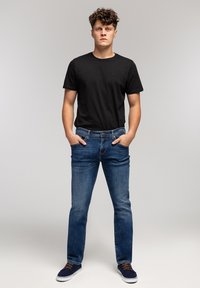 Czarna koszulka z okrągłym dekoltem, niebieskie jeansy, oraz granatowe trampki z brązowymi sznurowadłami. Model stoi prosto z rękami w kieszeniach, na neutralnym tle.