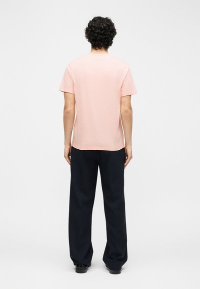 Άνδρας με κατσαρά μαλλιά που φοράει ανοιχτό ροζ κοντομάνικη μπλούζα και φαρδιά μαύρη παντελόνια, στέκεται γυρισμένος προς τα πίσω σε λευκό απλό φόντο.