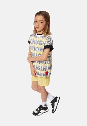 Camiseta de manga corta con un gráfico de texto amarillo y gris, y puños negros. Combinada con pantalones cortos amarillos claros y zapatillas en blanco y negro.