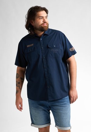 Marineblauw overhemd met korte mouwen en knopen, voorzien van twee borstzakken, een getextureerde logopatch en knoopdetails, gecombineerd met opgerolde denimshorts.
