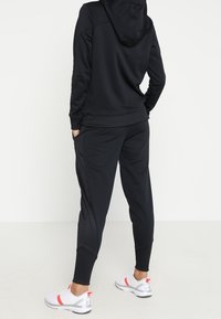 Felpa nera con cappuccio e pannelli testurizzati a contrasto, abbinata a pantaloni jogger coordinati. Calzature comprendono sneakers bianche con accenti rossi.