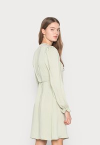 Vestido verde claro feito de tecido suave, com mangas longas bufantes, corpo ajustado e saia evasé com suave franzido na cintura.