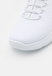 White athletic shoe with a textured mesh upper, featuring a rounded toe, elastic laces, and a rubber sole with subtle tread patterns.
