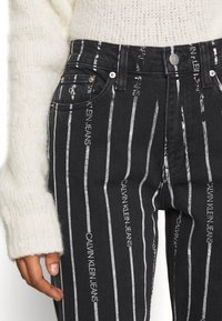 Svarta denimjeans med vertikala vita ränder och broderad text "Calvin Klein Jeans", utrustade med metallhardware och en figursydd design.