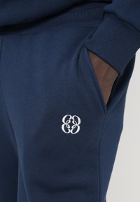 Navy blue sweatpants made of cotton blend fabric. Features side pockets, ribbed cuffs, and a white embroidered logo on the thigh.