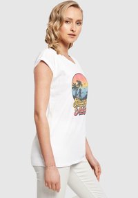Hvit kortermet t-skjorte med et trykk som viser en solnedgang, palmer og "Stranger Things"-tekst i gult og rødt. Glatt tekstur.