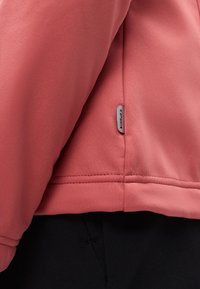 Gros plan sur l'ourlet d'une veste couleur saumon avec une petite étiquette de marque, portée sur un pantalon noir.