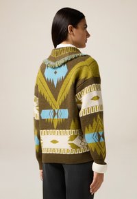 Neulepusero oliivinvihreänä, jossa on geometrisia kuvioita valkoisena ja vaaleansinisenä. Hapsullinen kaulus ja ribbireunus.