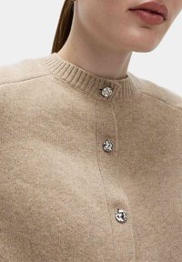 Beige stickad tröja med ribbad krage och tre dekorativa silverknappar längs fram. Mjuk textur och åtsittande design.