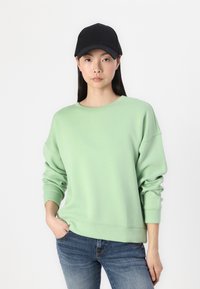 Sweatshirt oversize vert clair à manches longues, col rond et poignets côtelés, porté avec un jean bleu et une casquette noire.