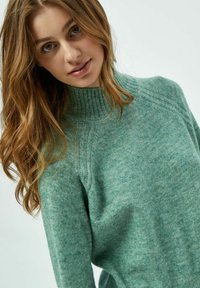 Maglione verde a maglia con collo alto, caratterizzato da dettagli a coste e una consistenza morbida, drappeggia comodamente sulle spalle.