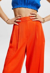 Světlé oranžové kalhoty s širokými nohavicemi, jemnou texturou a plisovaným předním dílem. Nosit vysokým pasem, v kombinaci s modrým krátkým topem a zlatými doplňky.