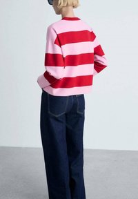 Pull en maille rayé rouge et rose avec un col rond, associé à un jean en denim bleu foncé à jambe large. Texture lisse, design décontracté.