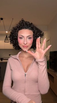 Femme aux cheveux bouclés portant un pull à fermeture éclair rose, levant une main avec cinq doigts visibles, debout dans une cuisine moderne.