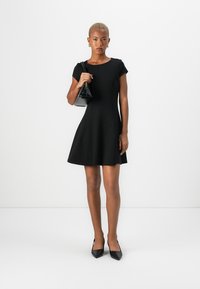 Robe noire ajustée avec une jupe évasée, des manches courtes et des coutures minimales. Le modèle tient un petit sac à main noir et porte des escarpins noirs à bout pointu.