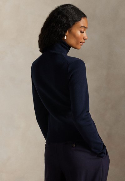 Γυναίκα με σκούρα σγουρά μαλλιά που φοράει μπλε σκούνο ζιβάγκο και ταιριαστό παντελόνι, στέκεται στο προφίλ με τα χέρια στις τσέπες.