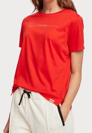 Mujer con camiseta roja de manga corta con texto blanco y pantalones con cordón color crema con ribete negro, con la mano en el bolsillo.