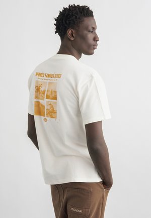 Mann iført hvit t-skjorte med gul vintage-stil "World Famous Dickies"-trykk på baksiden og brune bukser med "Pegador"-logo på lommen.