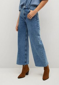 Jean denim bleu à jambes larges avec ourlet effiloché, taille haute et poches avant, associé à des bottines en suède marron.