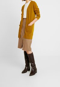Senfgelber Cardigan mit langen Ärmeln und zwei vorderen Taschen, kombiniert mit einem braun-gestreiften Rock in Karo-Optik. Braune, kniehohe, spitze Stiefel.
