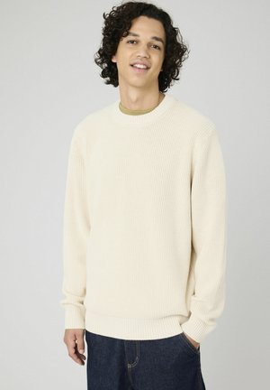Pull en maille côtelée couleur crème avec un col rond et des manches longues ; associé à un jean en denim foncé. Texture douce, coupe décontractée.