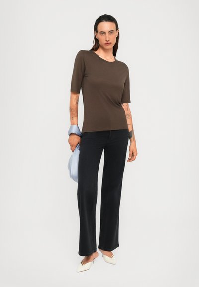 Woman with tattoos wearing a brown elbow-sleeve top, black pants, white pointed heels, holding a light blue bag against a white background.