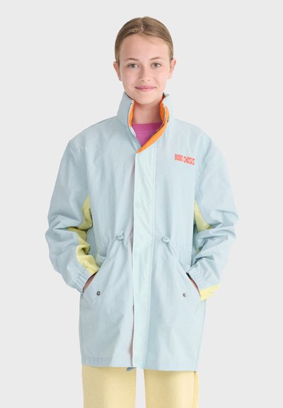 Jeune personne portant une veste bleu clair et jaune avec une doublure de fermeture éclair orange, debout les mains dans les poches devant un fond uni.