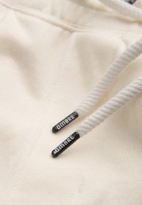 Gros plan des cordons blancs d'un sweat à capuche avec embouts noirs étiquetés "OMBRE" sur un fond en tissu beige doux.