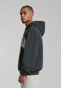 Grå hættetrøje med frontlomme, der har stor "MAN" tekst i en kontrastfarve. Blødt stof og løst fit design.