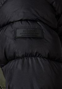 Pohled zblízka na černou vycpanou bundu se šitým štítkem, na kterém je napsáno "minoti Založeno v roce 1997 Navrženo ve Velké Británii." na zadní straně.