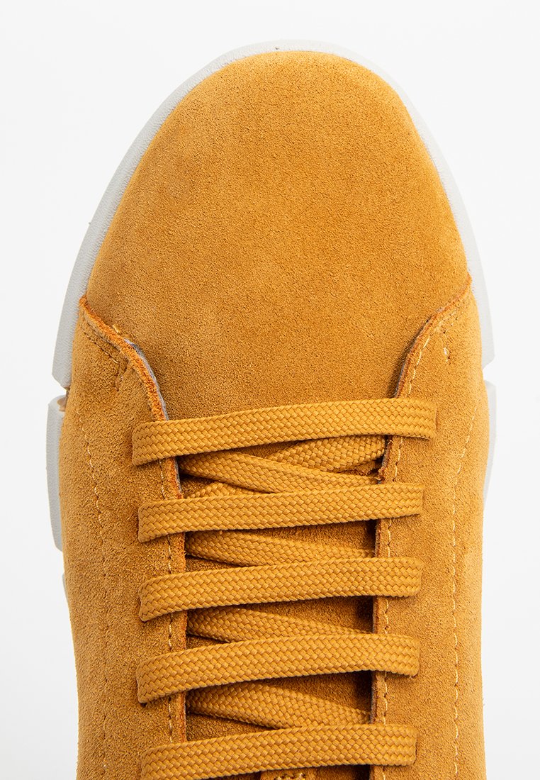 Żółte zamszowe sneakersy z płaskimi sznurowadłami, wyściółką na języku i białą gumową podeszwą. Nos ma zaokrąglony kształt i gładką fakturę.