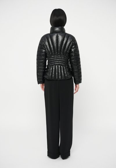 Doudoune noire avec un fini texturé et brillant, présentant une taille cintrée et un col haut ; portée avec un pantalon noir à jambes larges.