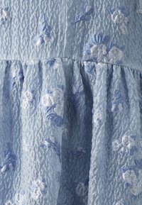 Tissu texturé bleu clair avec des détails plissés. Présente des motifs floraux brodés blancs et bleus sur toute sa surface.