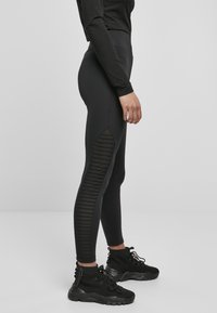 Leggings noirs taille haute avec des panneaux en maille côtelée sur les côtés, fabriqués dans un matériau extensible. Associés à des chaussures de sport noires à lacets.