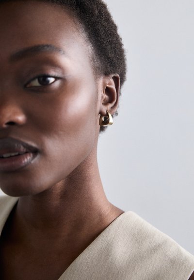 Gul geometrisk ørering med et buet design, båret af en model med kort, mørkt hår. Baggrunden er neutral og minimalistik.