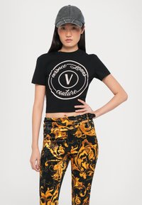 Camiseta negra corta con un logo crema, combinada con pantalones de terciopelo negros y dorados con un patrón, de talle alto y cinturón decorativo.
