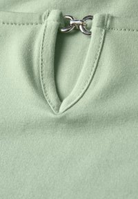 Ljusgrön topp med en texturerad, ribbad tyg. Har en nyckelhålsformad halsringning säkrad med en silverfärgad ögla som accent.