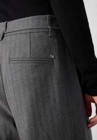 Gros plan d'un pantalon gris en chevrons avec une poche passepoilée arrière et un petit badge de marque argenté près de la poche, porté avec un haut noir.