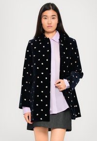 Frau, die einen navyblauen, blumenbedruckten Samtmantel über einem helllila Hemd und einem dunkelgrauen Rock trägt, steht vor einem schlichten Hintergrund.