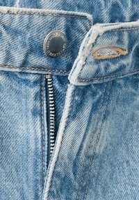 Jean en denim bleu clair avec fermeture éclair métallique partiellement ouverte et bouton visible étiqueté "KING DENIM."