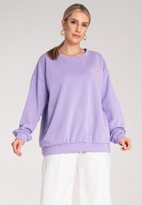 Fioletowy, oversized sweatshirt z ryflowanymi mankietami i paskiem w talii; posiada haft na przodzie w kontrastującym różowym kolorze; miękki, gładki w dotyku.