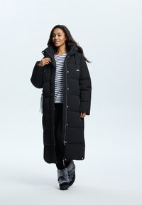 Manteau d'hiver long noir avec capuche, présentant un design matelassé, une fermeture éclair métallique et des cordons de serrage réglables, porté sur un haut rayé et un pantalon noir.