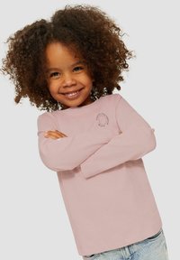 Lachend kind met krullend haar, gekleed in een roze lange mouw shirt en lichtblauwe jeans, met vertrouwen de armen gekruist tegen een witte achtergrond.